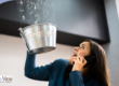 Woman holding a bucket to catch water leaking from the ceiling while on the phone.